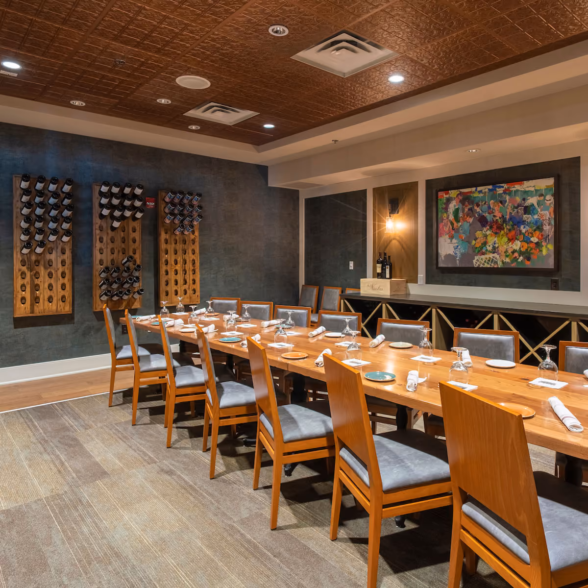 Private dining room with a long wooden table set with plates, napkins, and upside-down wine glasses, surrounded by wooden chairs with gray cushions, wine racks on the wall, and a colorful painting.