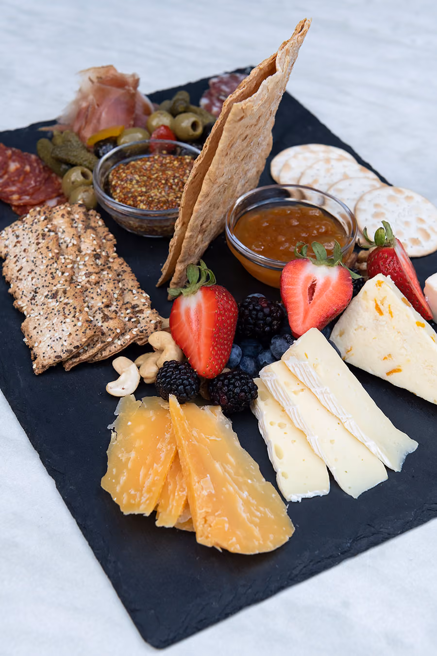 Cheese board with assorted cheeses, crackers, strawberries, blackberries, nuts, cured meats, pickles, olives, grain mustard, and apricot jam.
