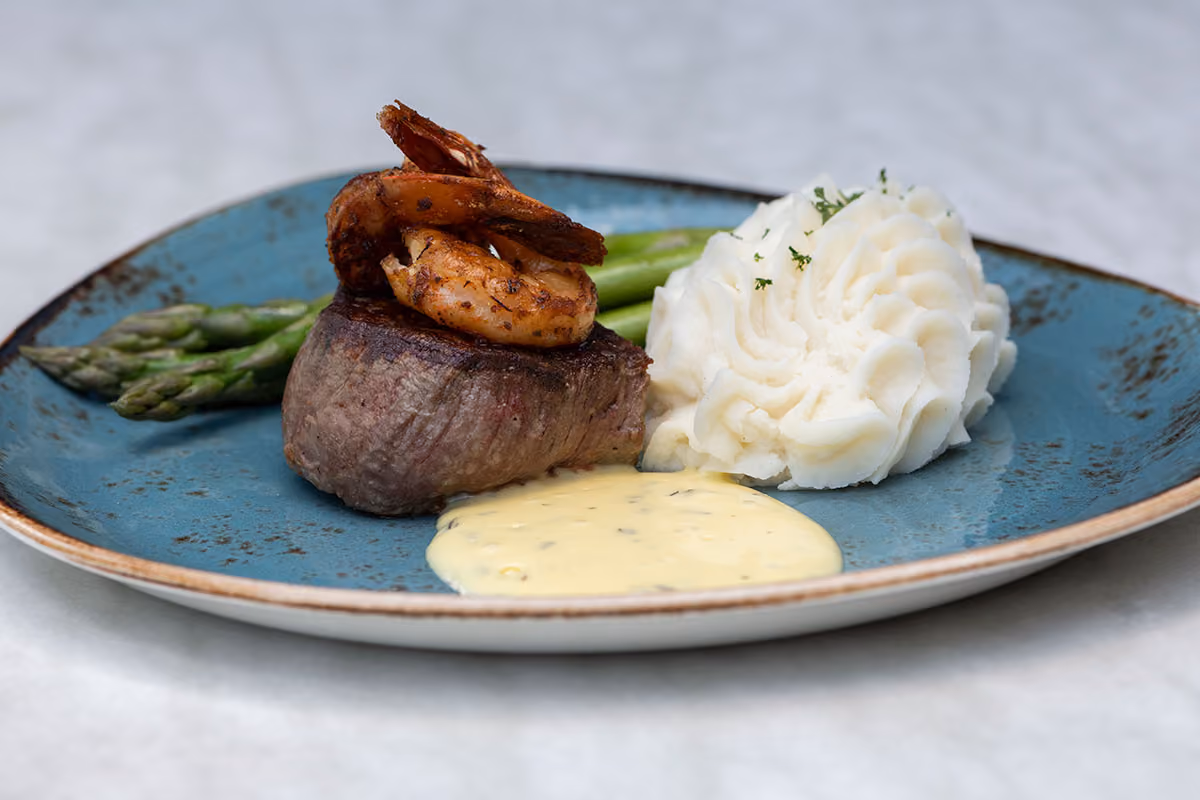 Grilled filet mignon topped with two seasoned shrimp, served with mashed potatoes, asparagus, and a creamy mustard sauce on a blue plate.