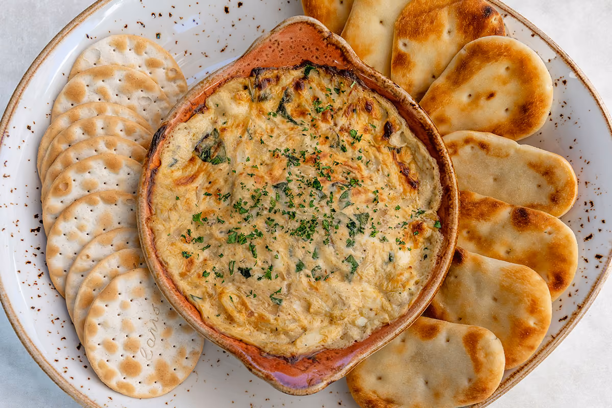 Baked creamy spinach and artichoke dip garnished with parsley, served with round and oval crackers on a white speckled plate.