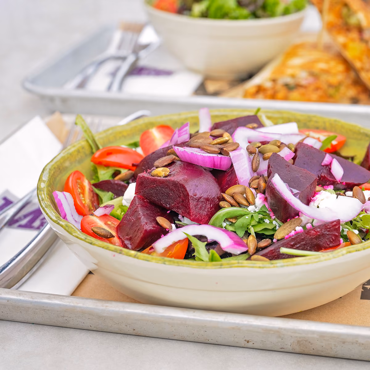 Bowl of salad with beet chunks, cherry tomatoes, red onion slices, pumpkin seeds, greens, and crumbled cheese.