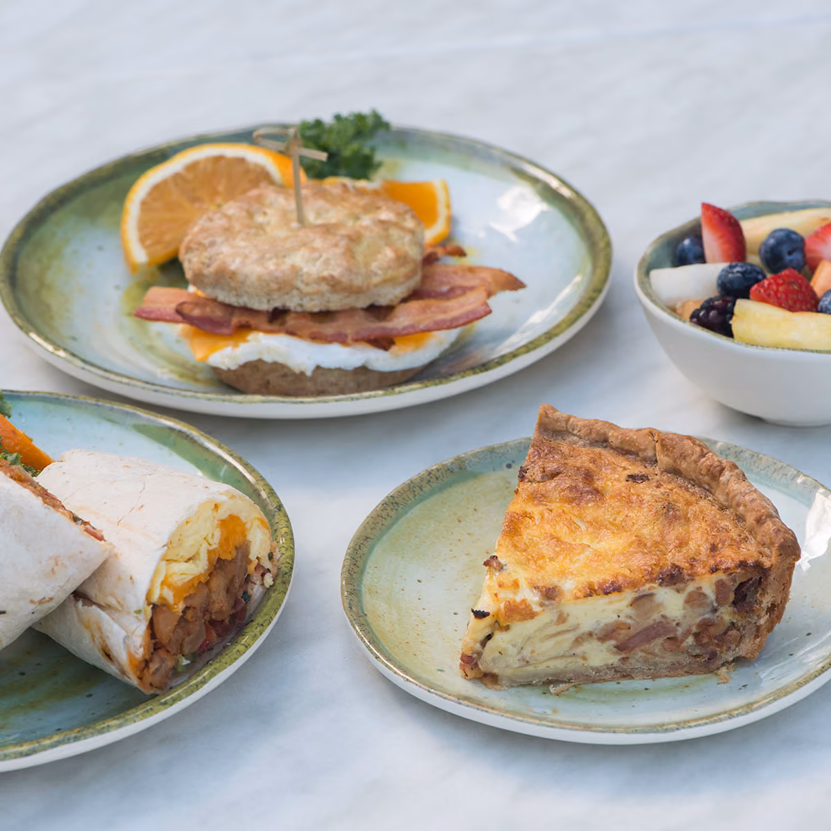 Breakfast plate with a bacon and egg biscuit sandwich, a breakfast burrito, a slice of quiche, and a bowl of mixed fruit.