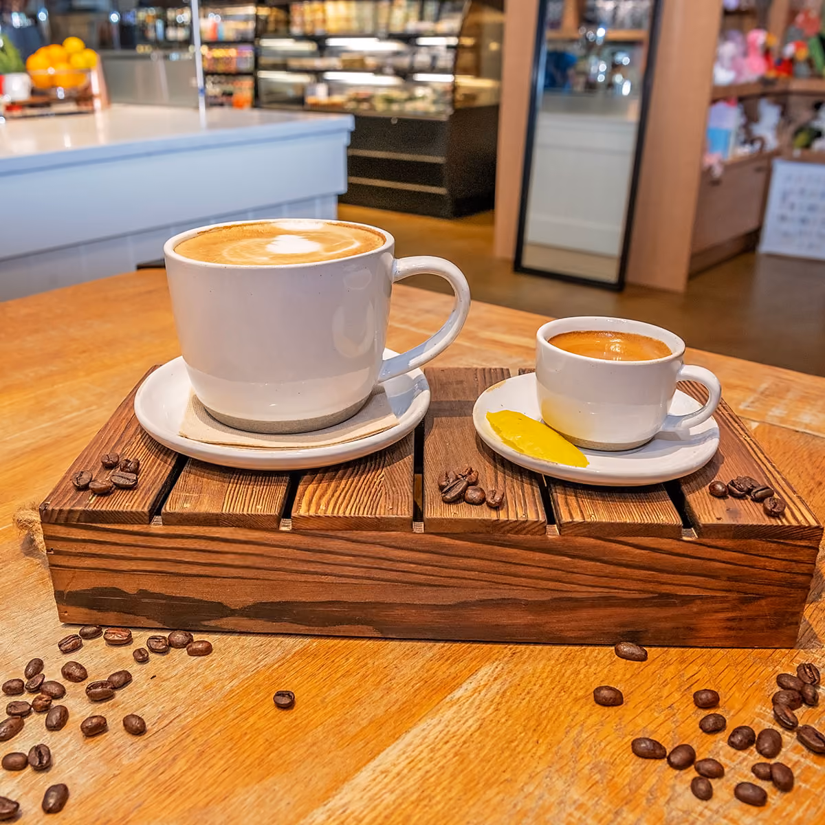 A large cup of latte with latte art and a small cup of espresso with a yellow garnish, served on a wooden tray with coffee beans scattered around.