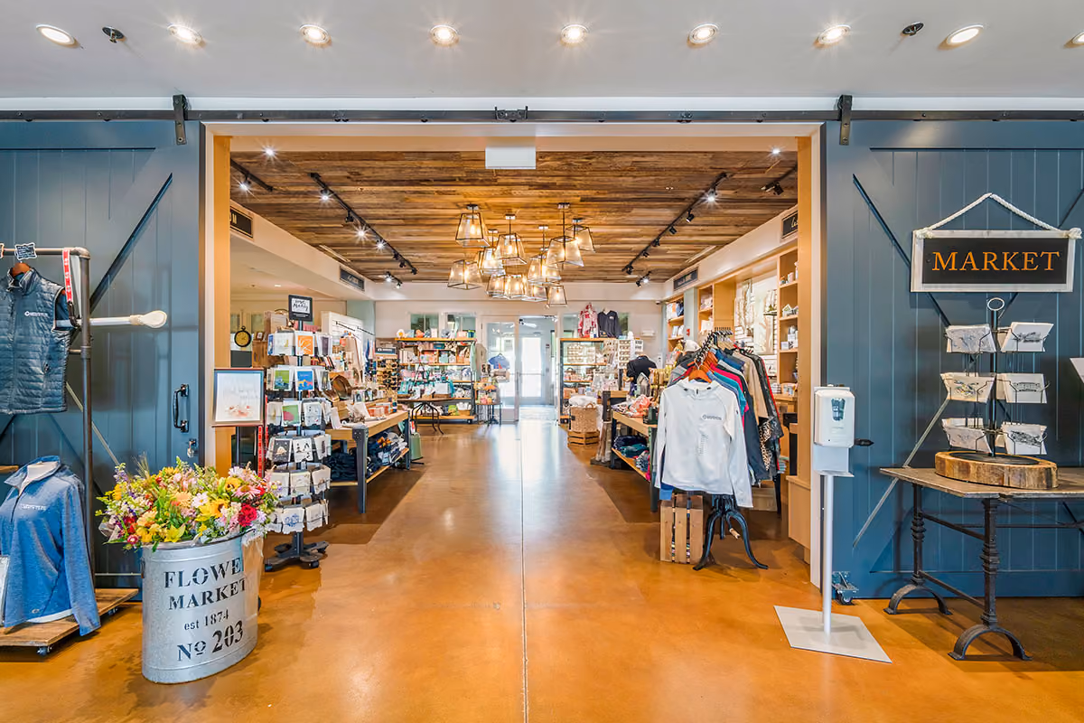 Interior view of a bright market store with clothing racks, flower bucket, display shelves, and decorative hanging lights.