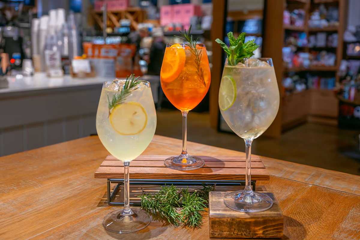 Three cocktails on a wooden table, each in a wine glass with ice; one garnished with lemon and rosemary, one with orange and rosemary, and one with lime and mint.