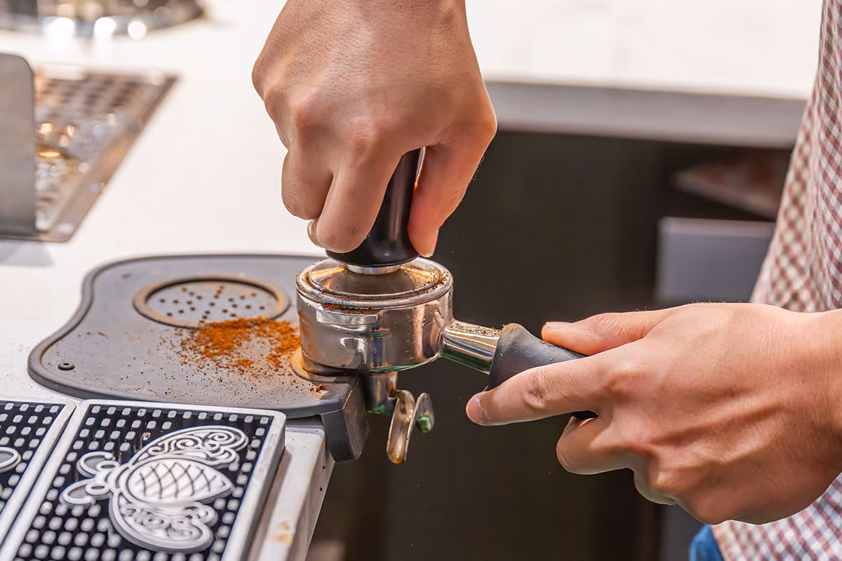 Barista tamping ground coffee in a portafilter on a coffee machine.