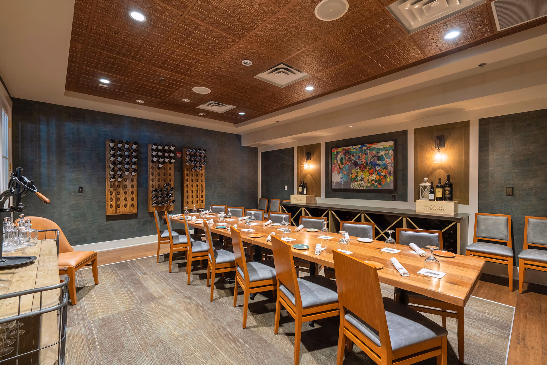 Elegant private dining room with a long wooden table set with plates, glasses, and napkins, surrounded by wooden chairs with gray cushions, wine racks on a dark wall, and decorative lighting.