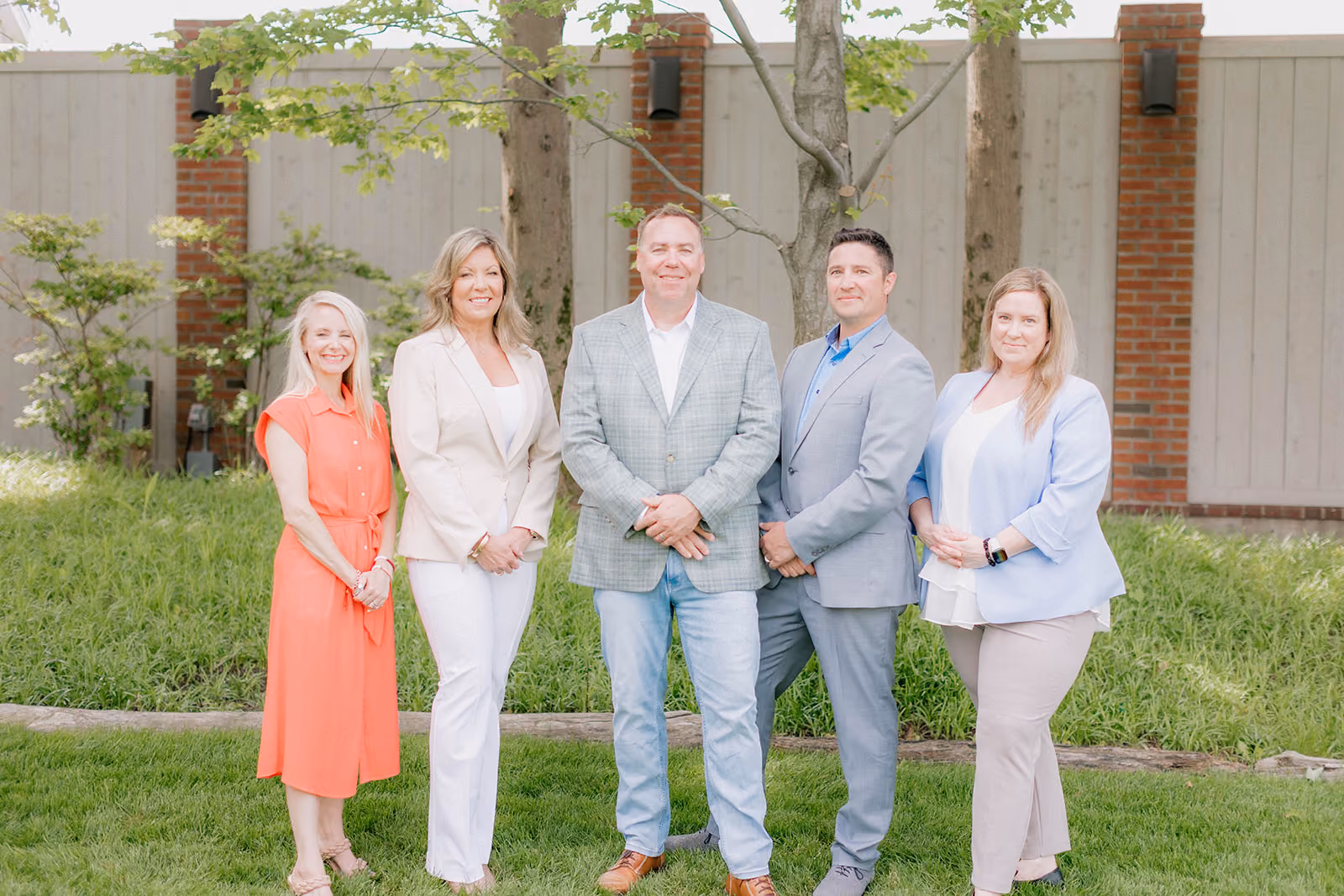 Five diverse professionals standing outdoors on grass in front of a wooden fence and trees, dressed in business casual attire and smiling.