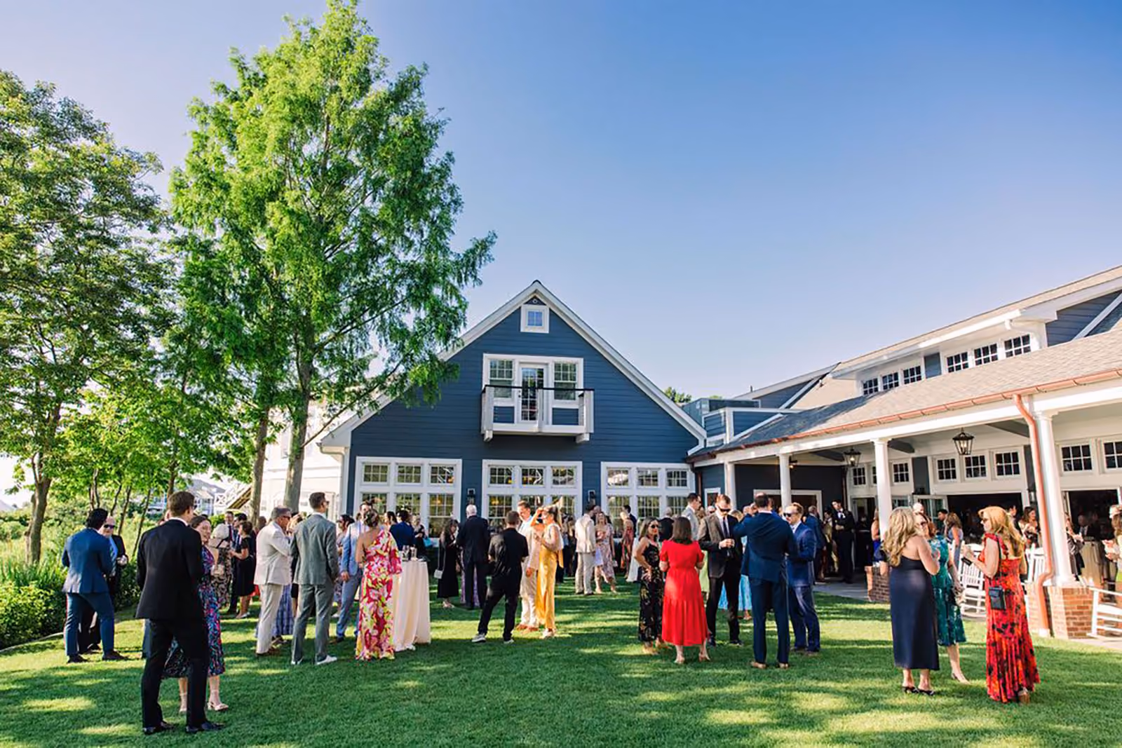 People dressed in formal attire socializing outdoors on a green lawn in front of blue and white buildings under a clear sky.