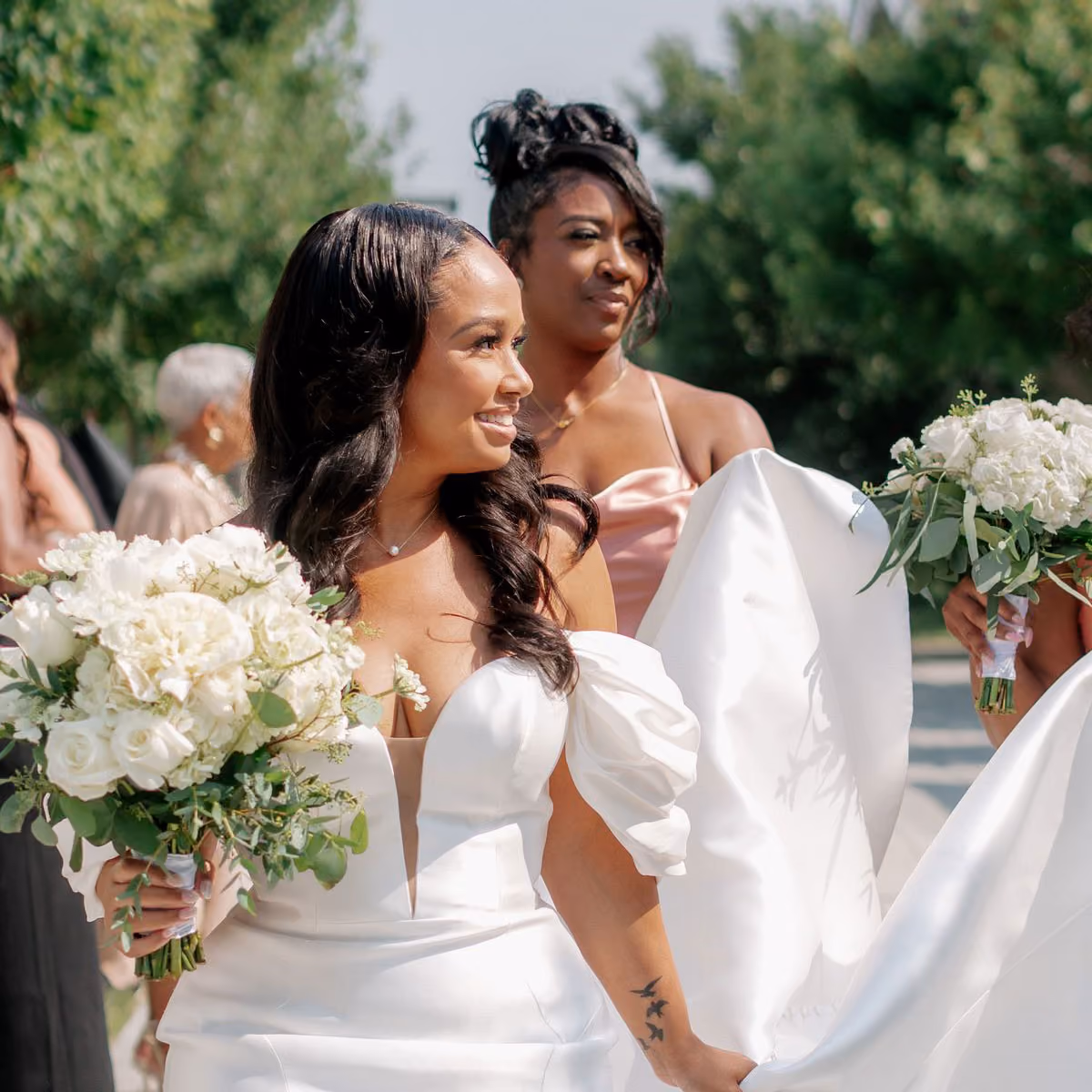 Smiling bride in a white wedding dress holding a bouquet of white flowers, accompanied by a bridesmaid in a pink dress holding the bride's gown.