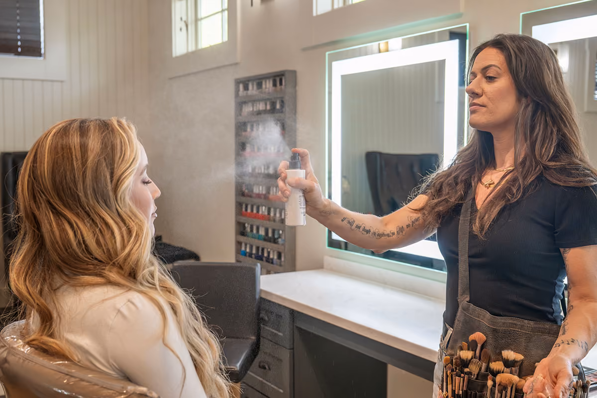 Hair stylist spraying hair spray on a seated client with wavy blonde hair in a bright salon.