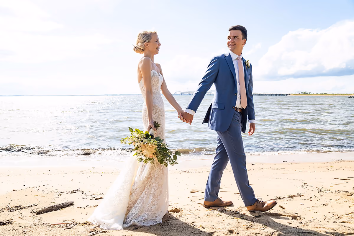 Bride and groom holding hands and walking on a sandy beach beside the water.