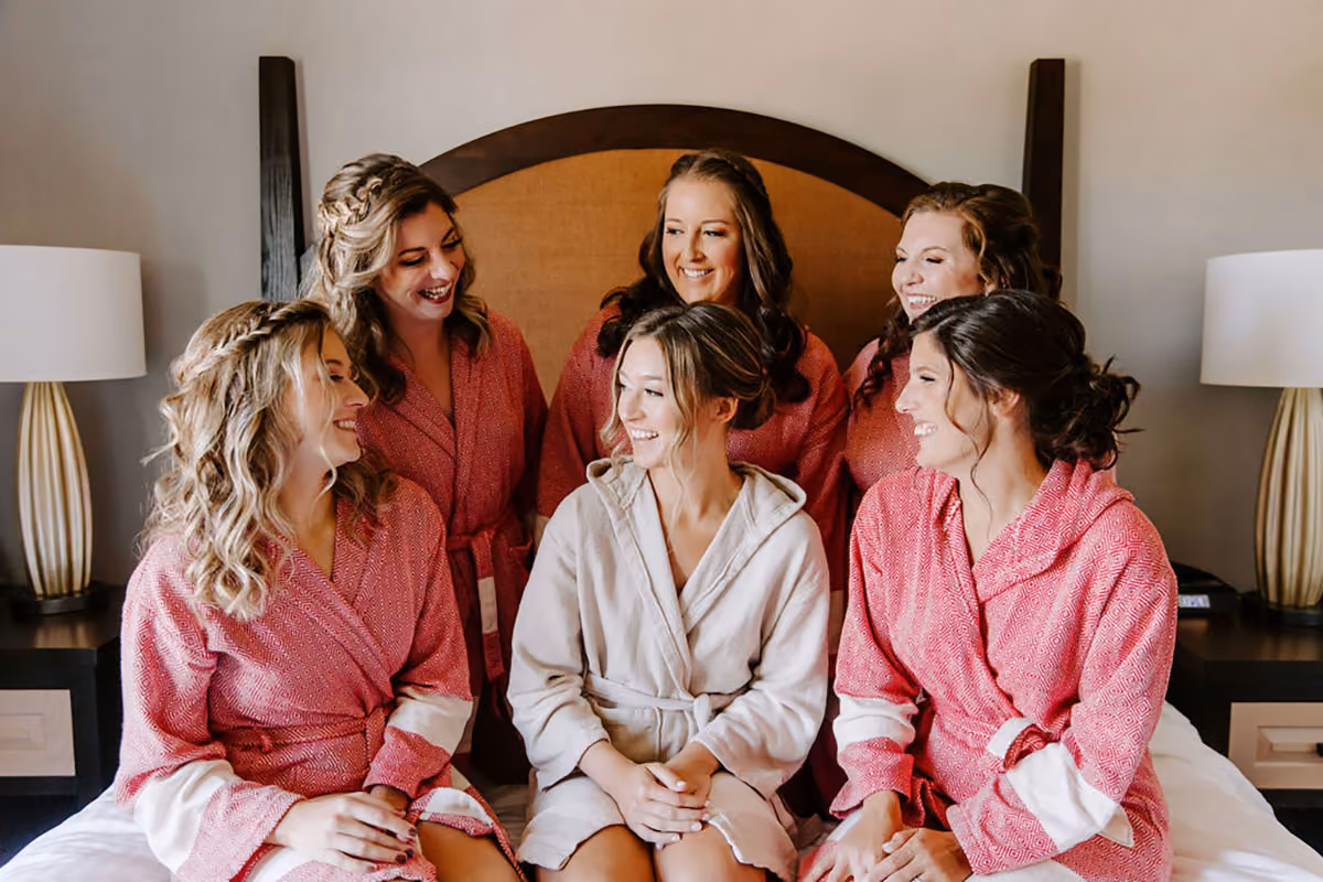 Bride in white robe smiling with five bridesmaids in matching pink robes sitting on a bed in a bedroom.