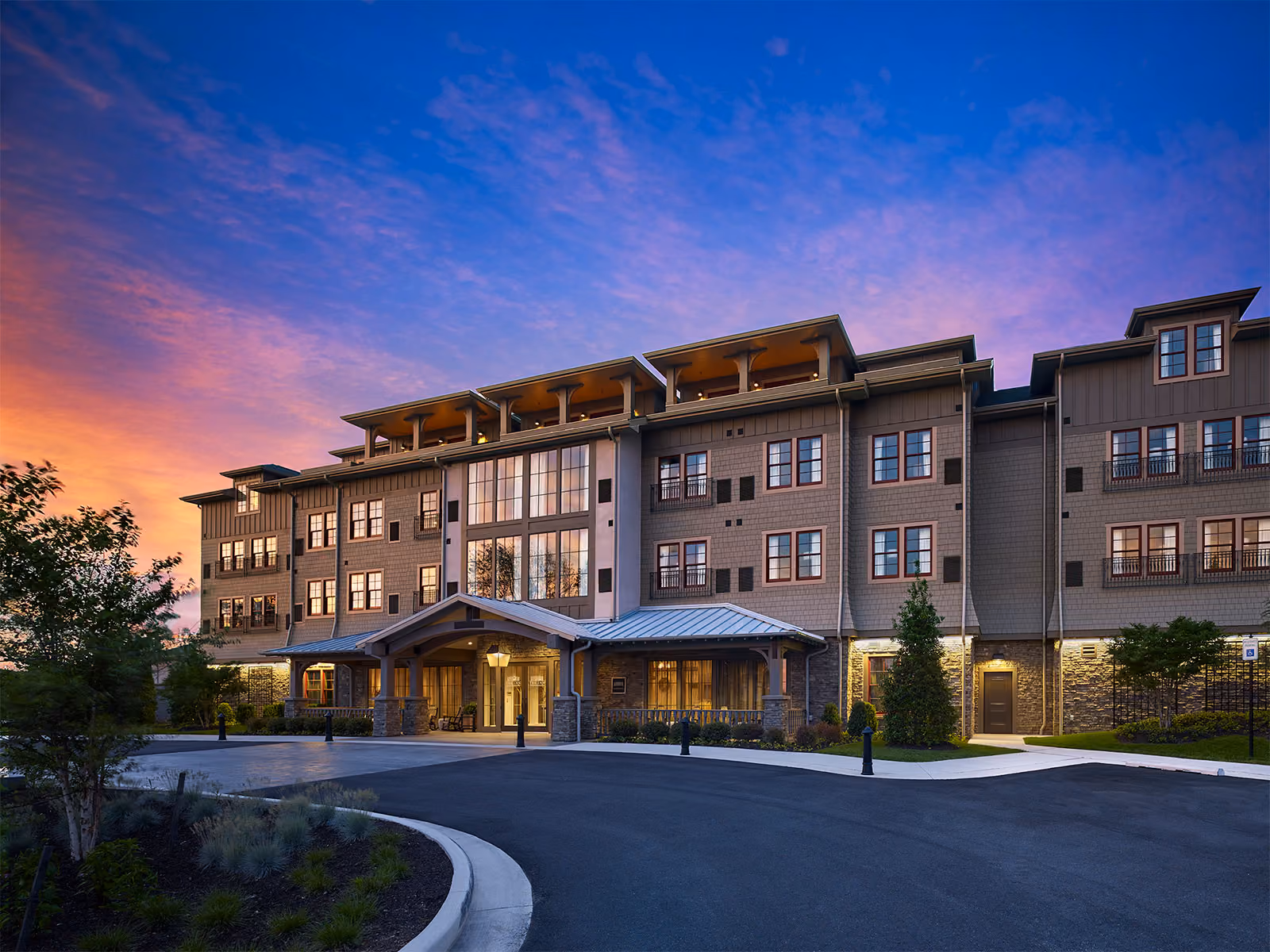 A modern four-story residential building with large windows, lit up at dusk under a colorful sky.