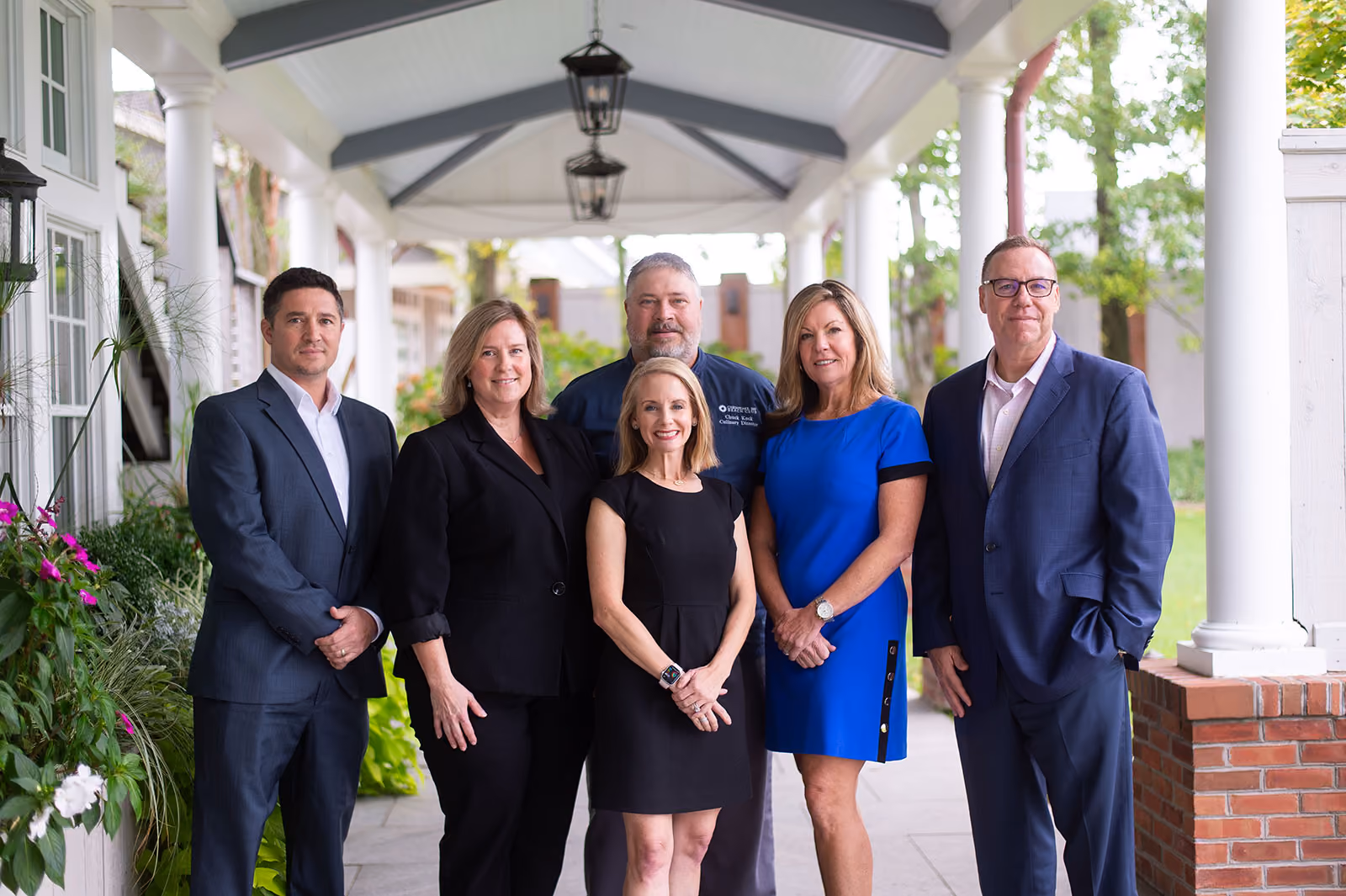 Six professionally dressed adults stand in a row under a covered outdoor walkway, posing for a group photo.