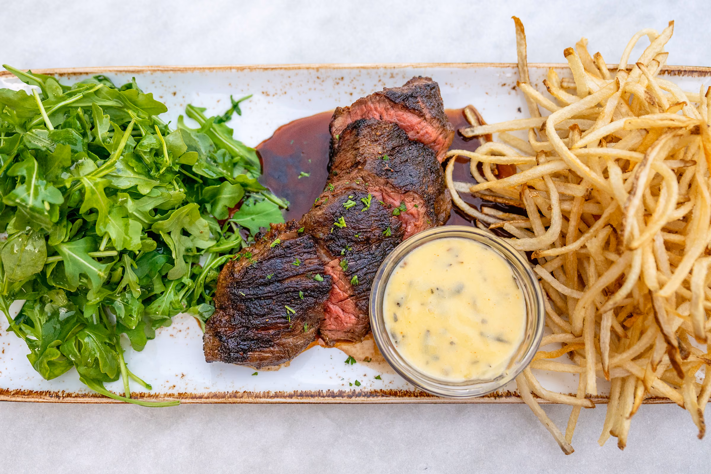 Grilled steak with sauce, arugula salad, and shoestring fries served on a rectangular plate.