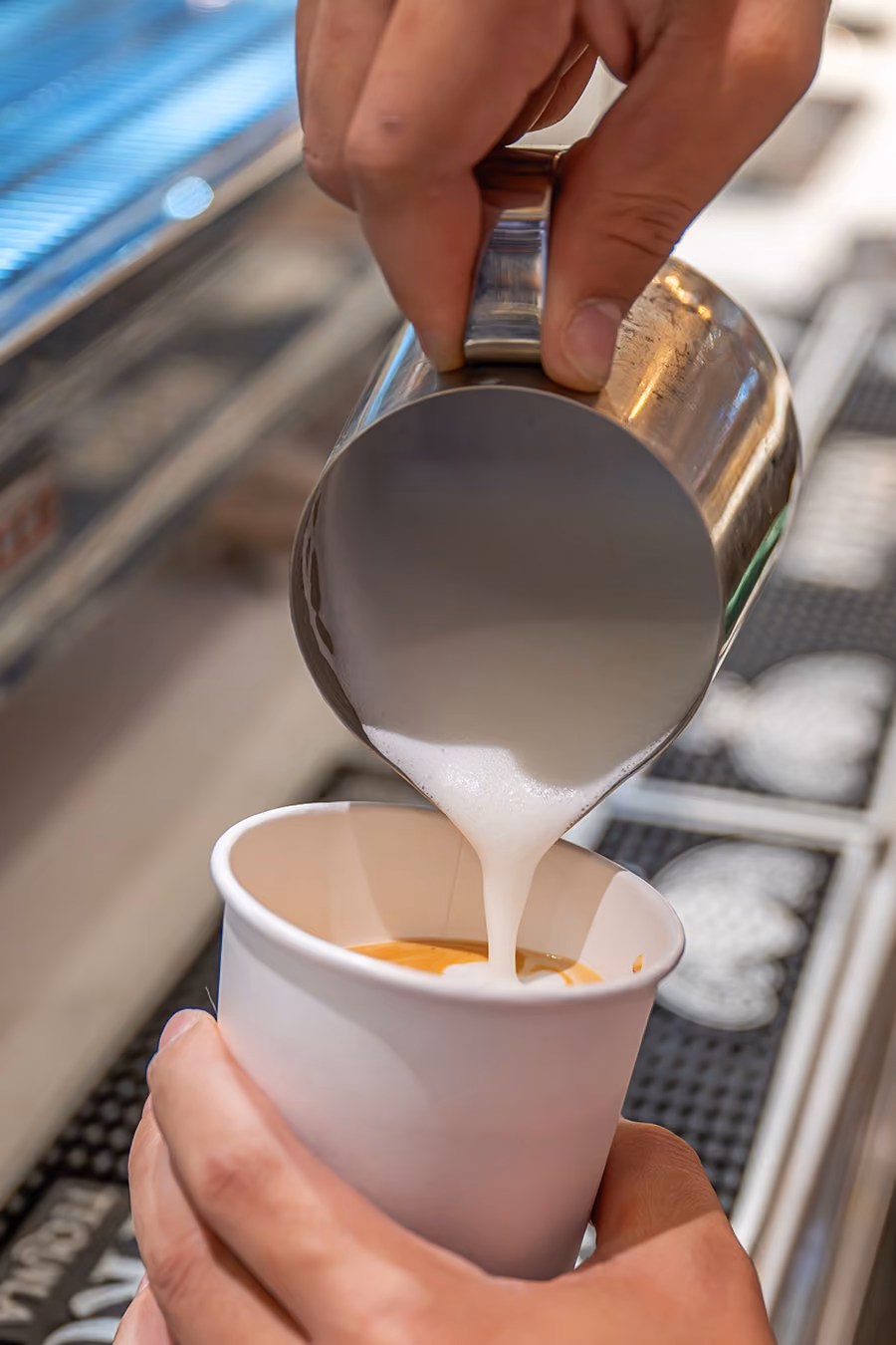 A person pours steamed milk from a metal pitcher into a paper cup of espresso at a coffee bar.