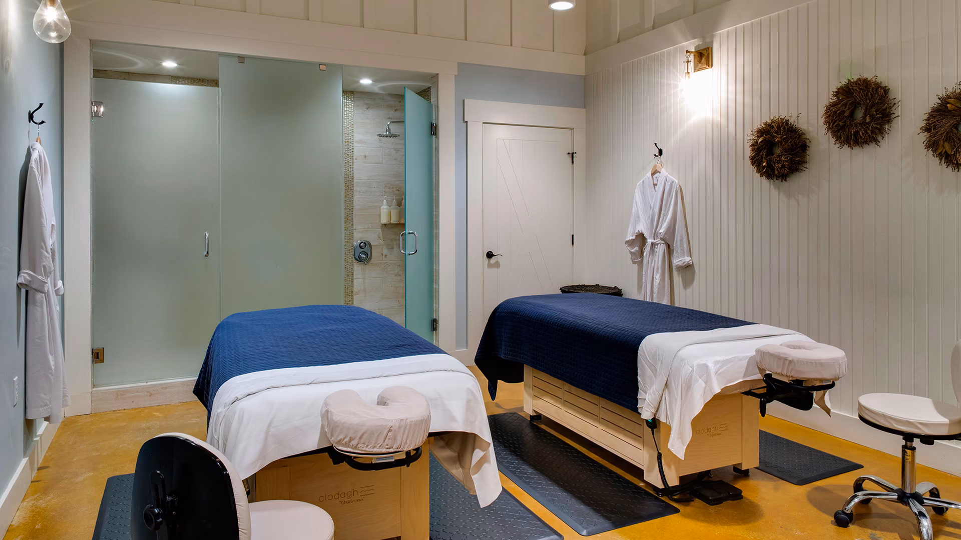 Two massage tables with navy covers in a spa room, white walls, robes hanging, and a glass shower in the background.