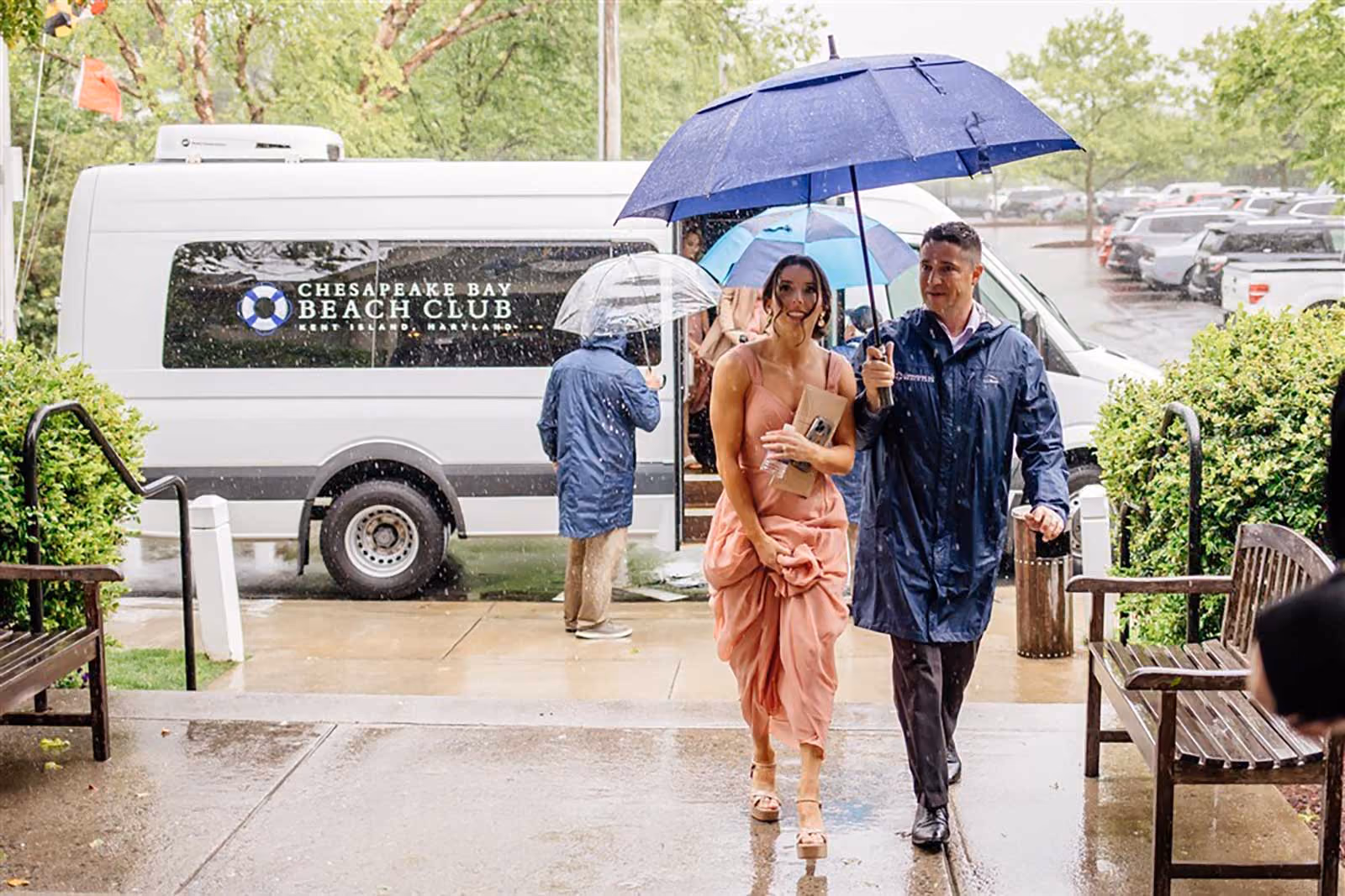 A woman in a pink dress and a man in a raincoat walk under an umbrella in the rain near a Chesapeake Bay Beach Club van.