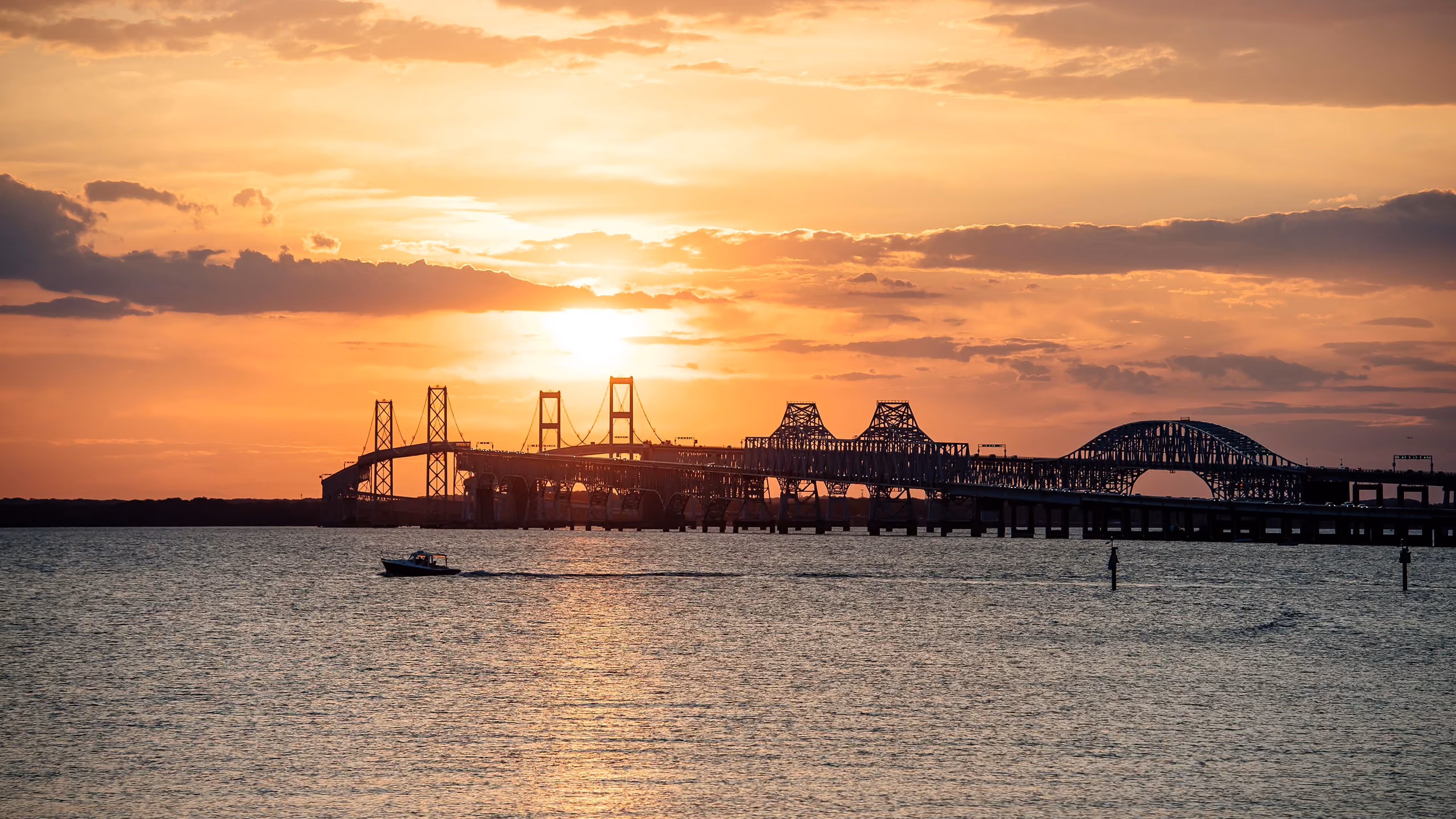 Boat on water near a long bridge at sunset with orange sky and clouds.