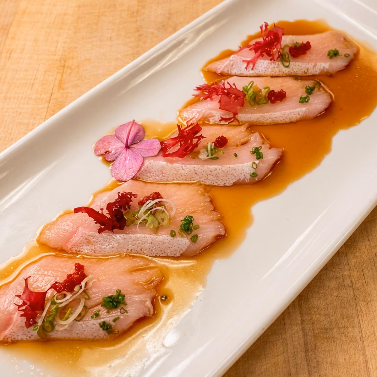 Yellowtail sashimi slices arranged with citrus ponzu, red seaweed, scallions and edible flowers on a rectangular plate at Vue