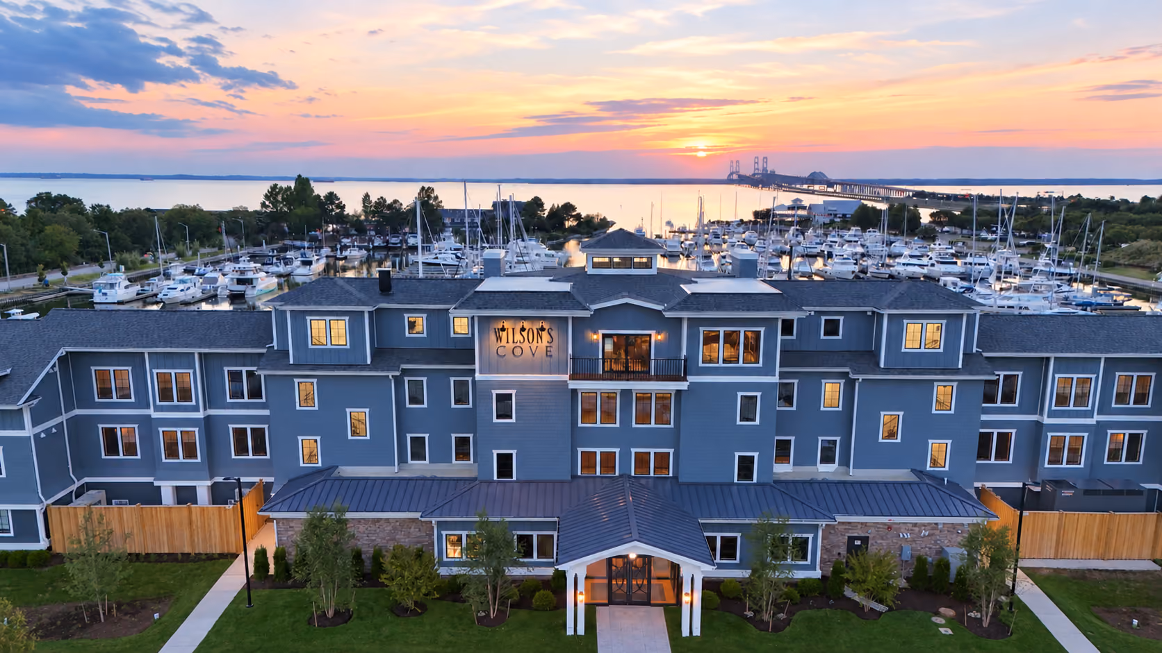 Gray multi-story building with marina and boats behind it, under a sunset sky by the water.