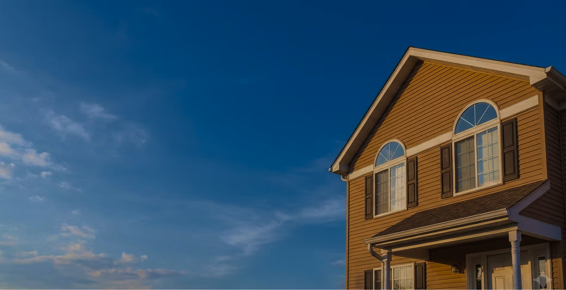 Upper portion of a beige two-story house with two arched windows and blue sky with scattered clouds.