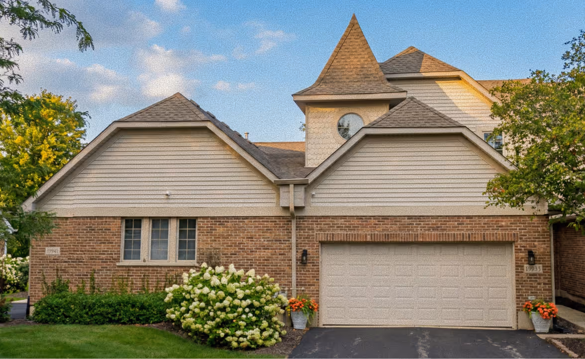 Suburban brick house with beige siding, a double garage door, a pointed turret, and flowering bushes in the front yard under a partly cloudy sky.