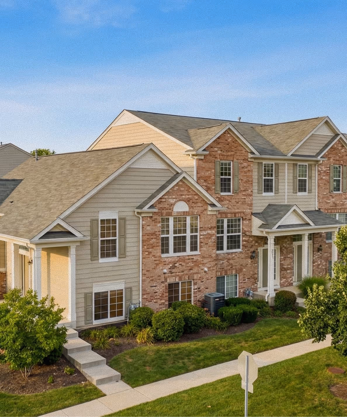 Two-story residential building with beige siding and red brick facade under a clear blue sky.
