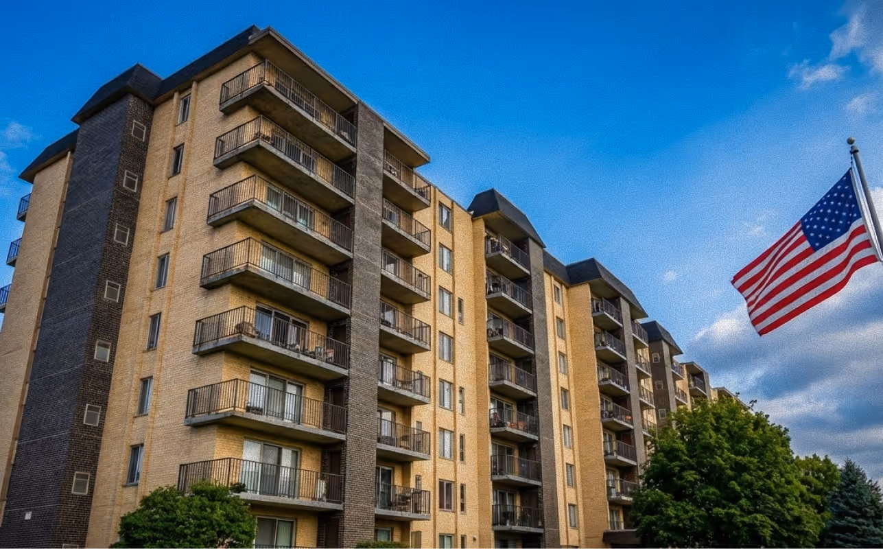 Multi-story residential building with balconies under a blue sky and an American flag on a flagpole nearby.