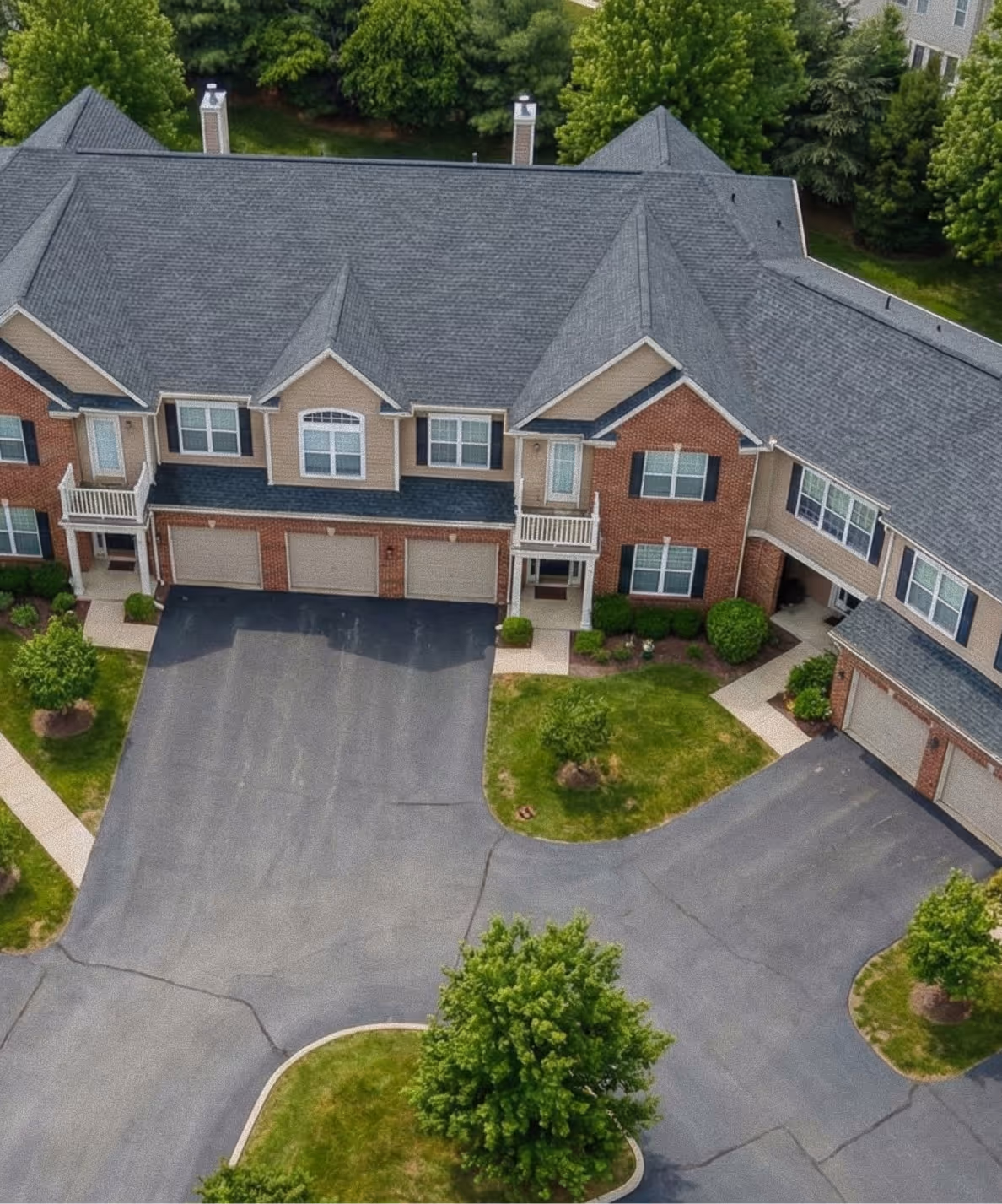 Aerial view of a residential townhouse complex with multiple garages, paved driveways, and surrounding green trees and lawns.