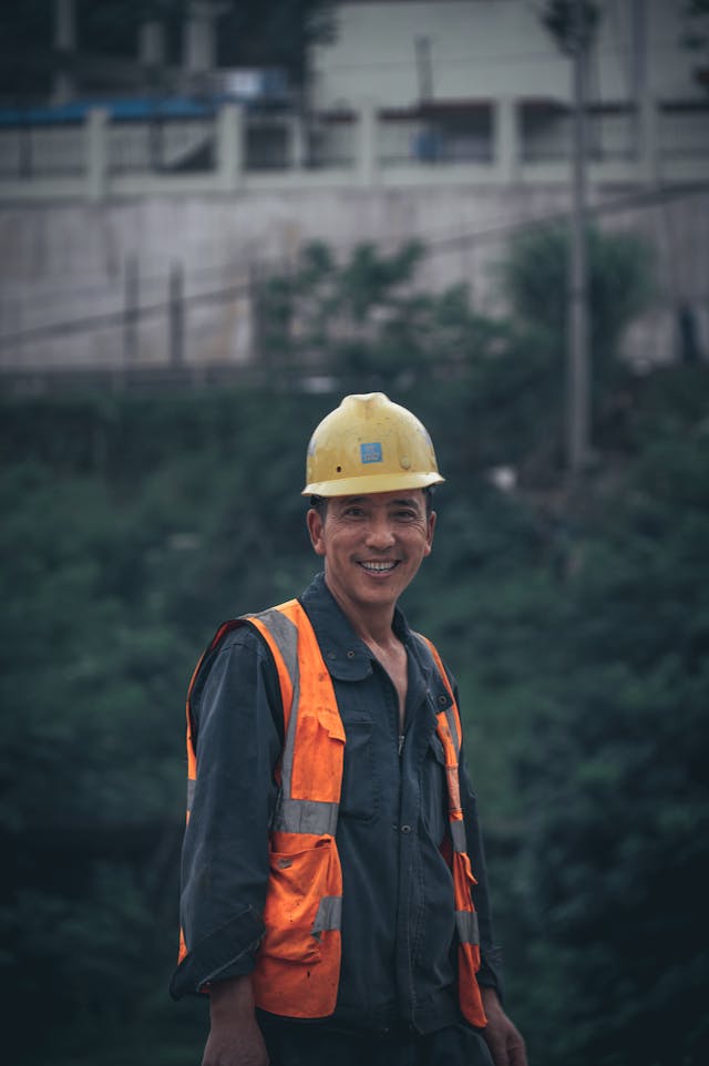 Smiling construction worker wearing a yellow hard hat and orange safety vest outdoors.