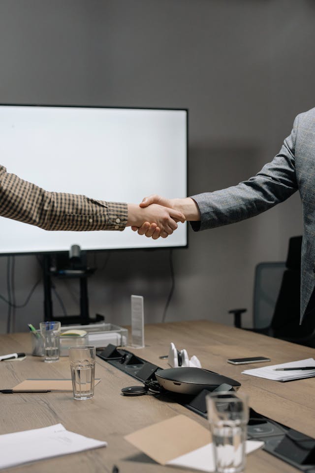 Two people shaking hands over a conference table with glasses and office supplies.