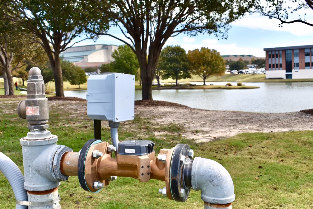 Water pipe infrastructure with a monitoring device near a pond in a park-like setting with trees and buildings in the background.