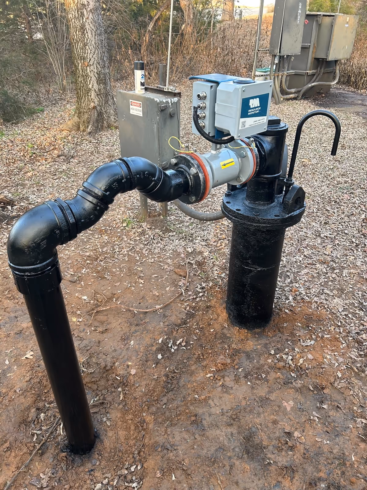 Outdoor black metal water meter pipe assembly with a digital resource monitor and electrical box in a wooded area.