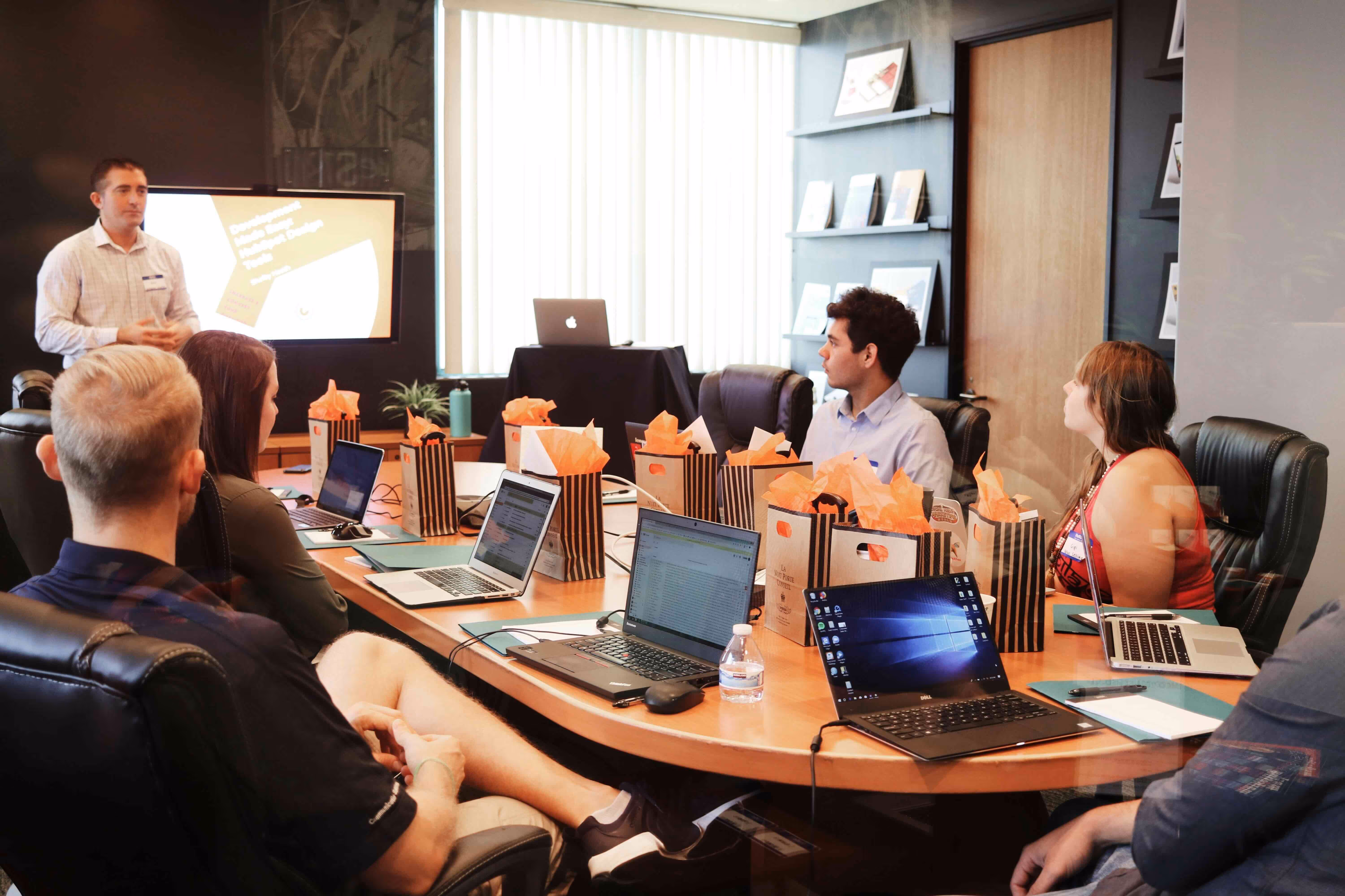 Group of people seated around a conference table watching a presentation with laptops and gift bags on the table.