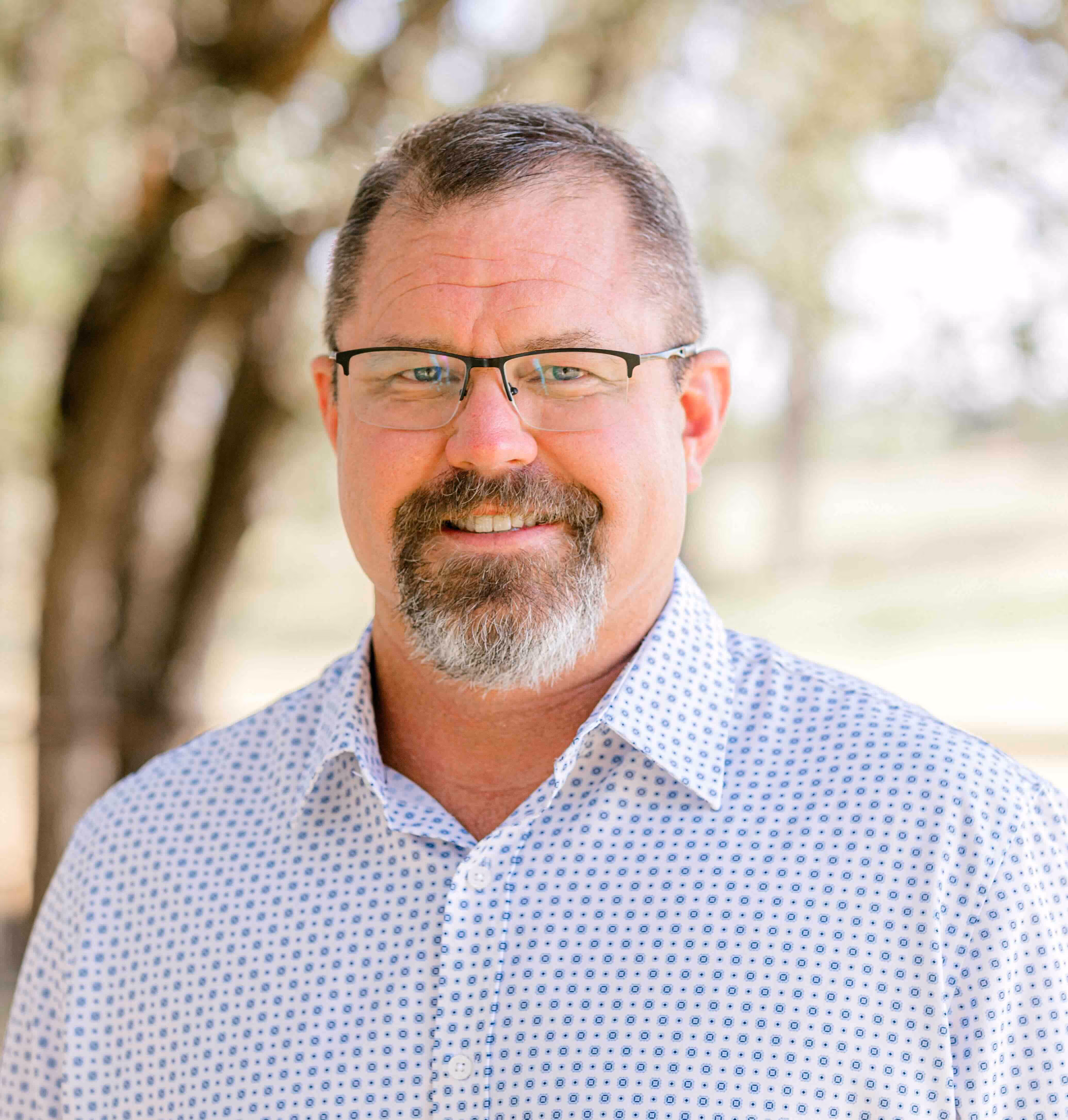 Middle-aged man with short hair, glasses, and a salt-and-pepper goatee wearing a white patterned button-up shirt, smiling against a blurred outdoor background.