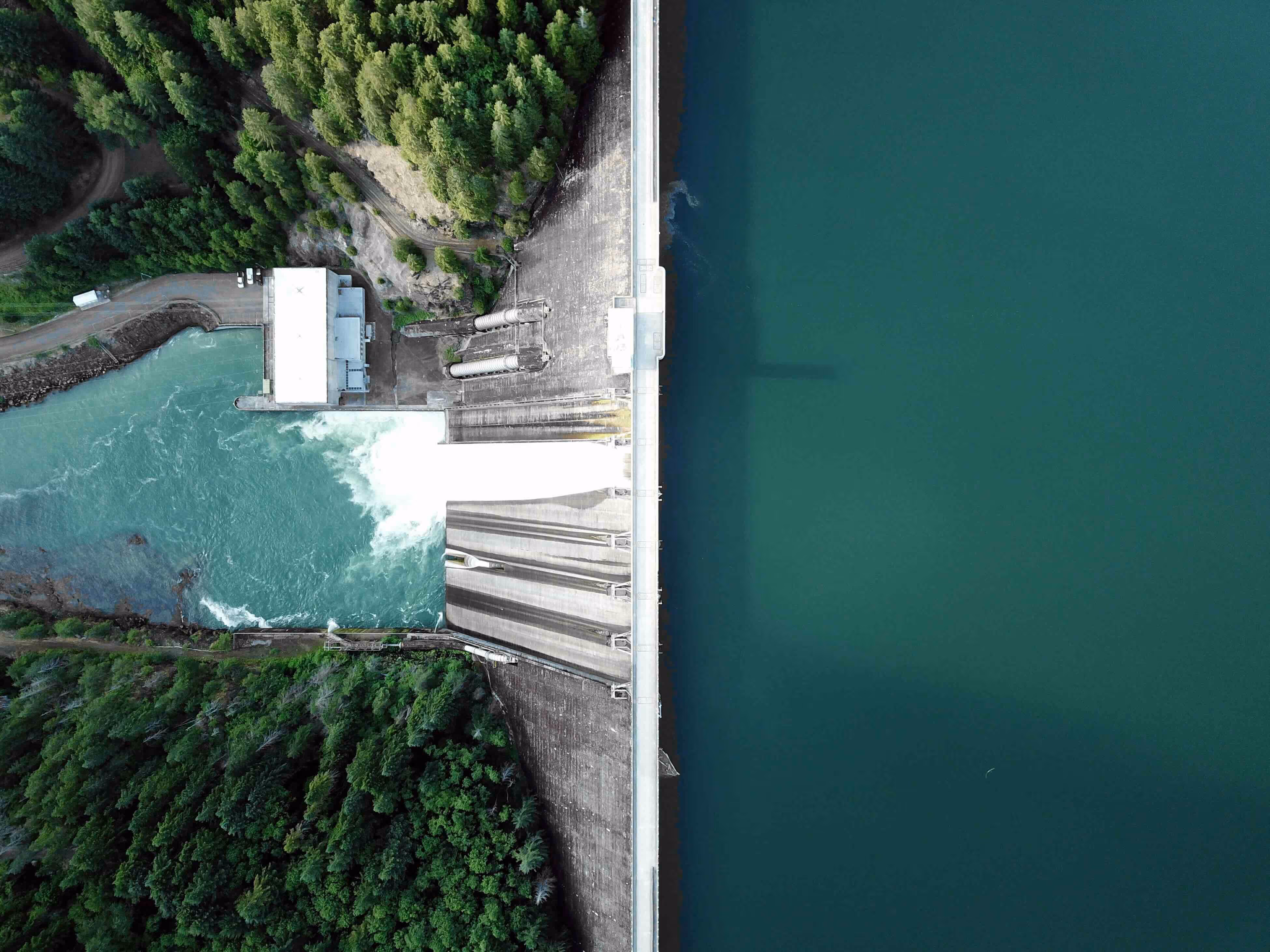 Aerial view of a dam with water flowing through the spillways, surrounded by dense green forest.