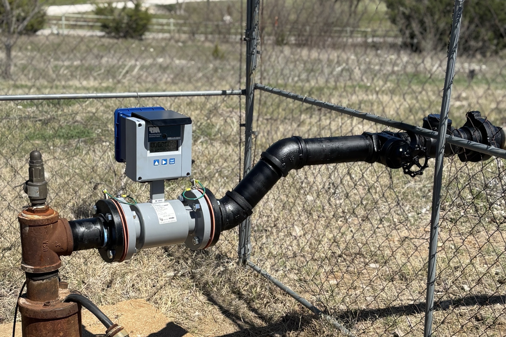 Flow meter with a digital display on a wellhead pipe inside a chain-link fence, with dry grass and open land behind.