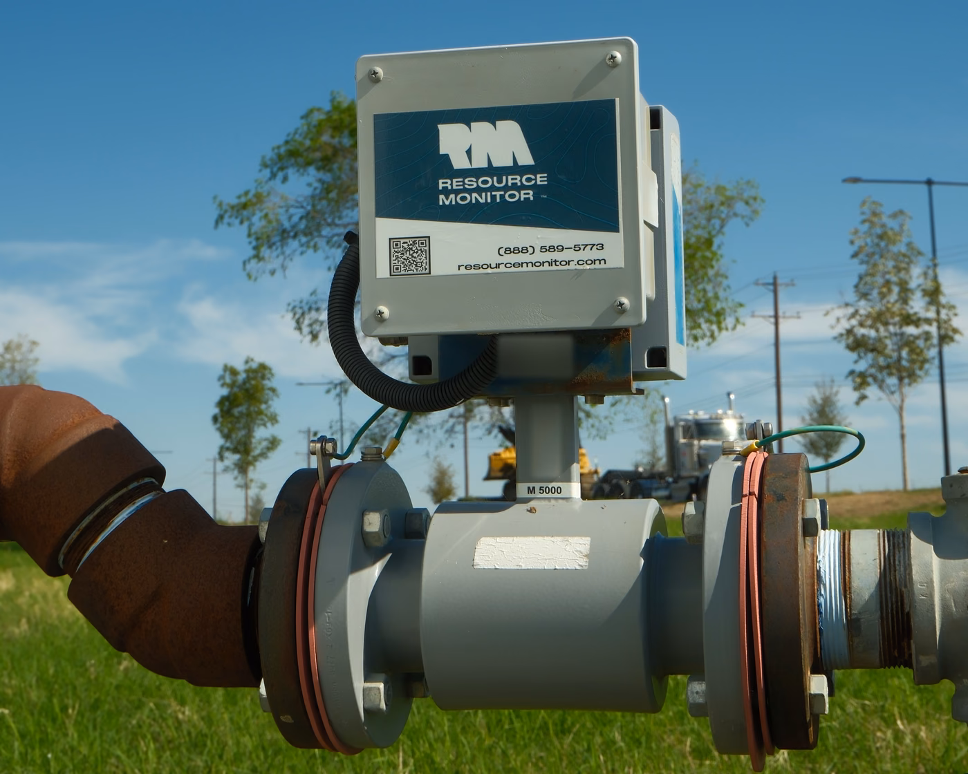 Resource Monitor M5000 flow meter installed on a water line in an open field, with the branded display box and QR code under a blue sky.