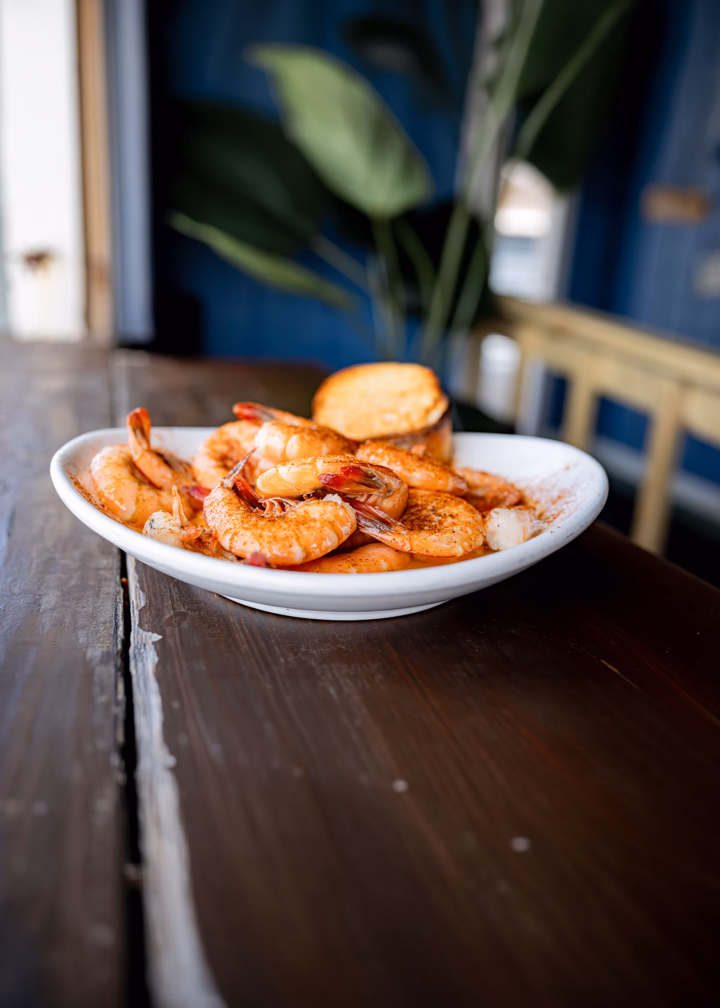 White plate with seasoned cooked shrimp and a piece of toasted bread on a wooden table with blurred background.