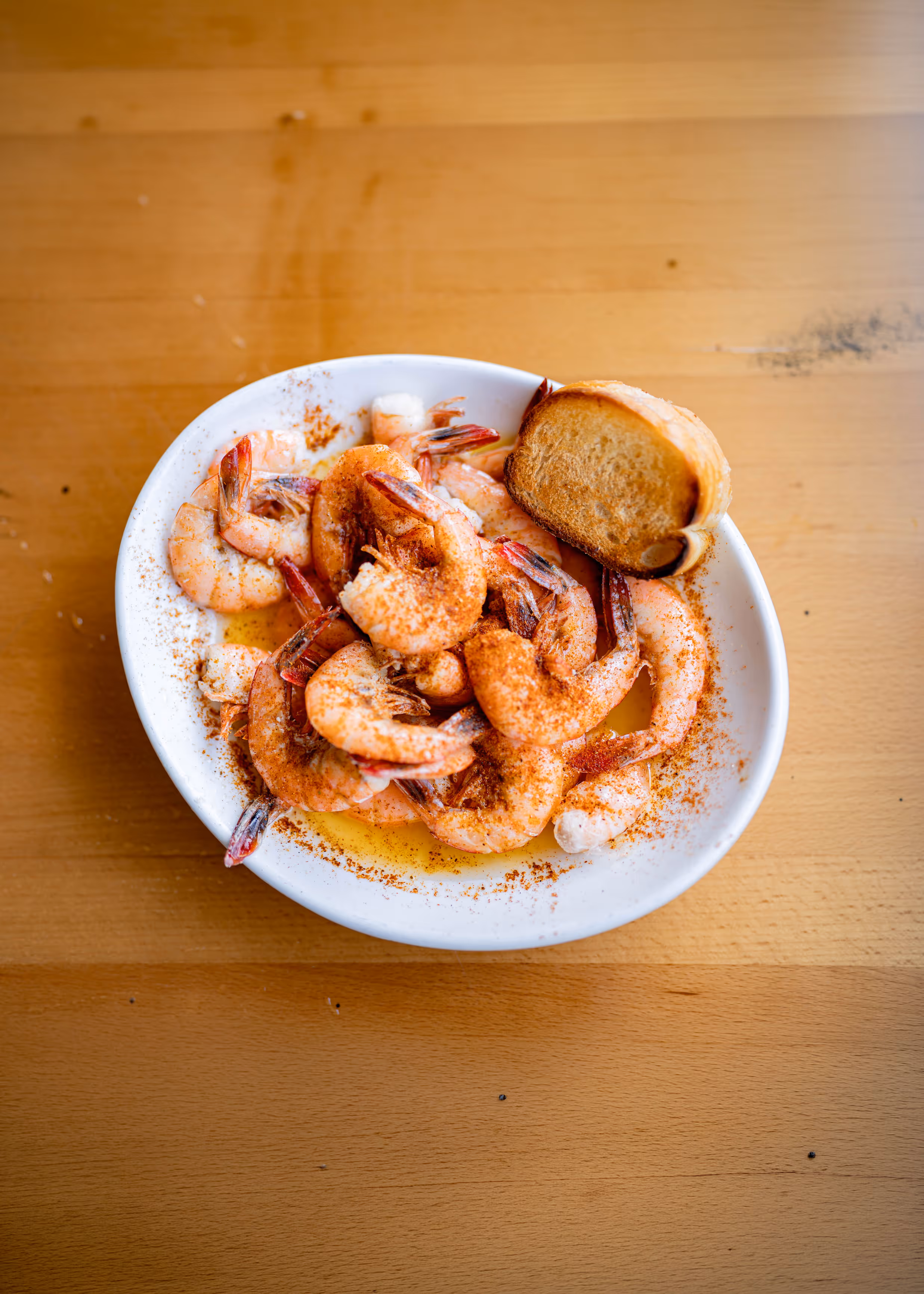 White bowl of cooked shrimp seasoned with spices and accompanied by a slice of toasted bread on a wooden table.