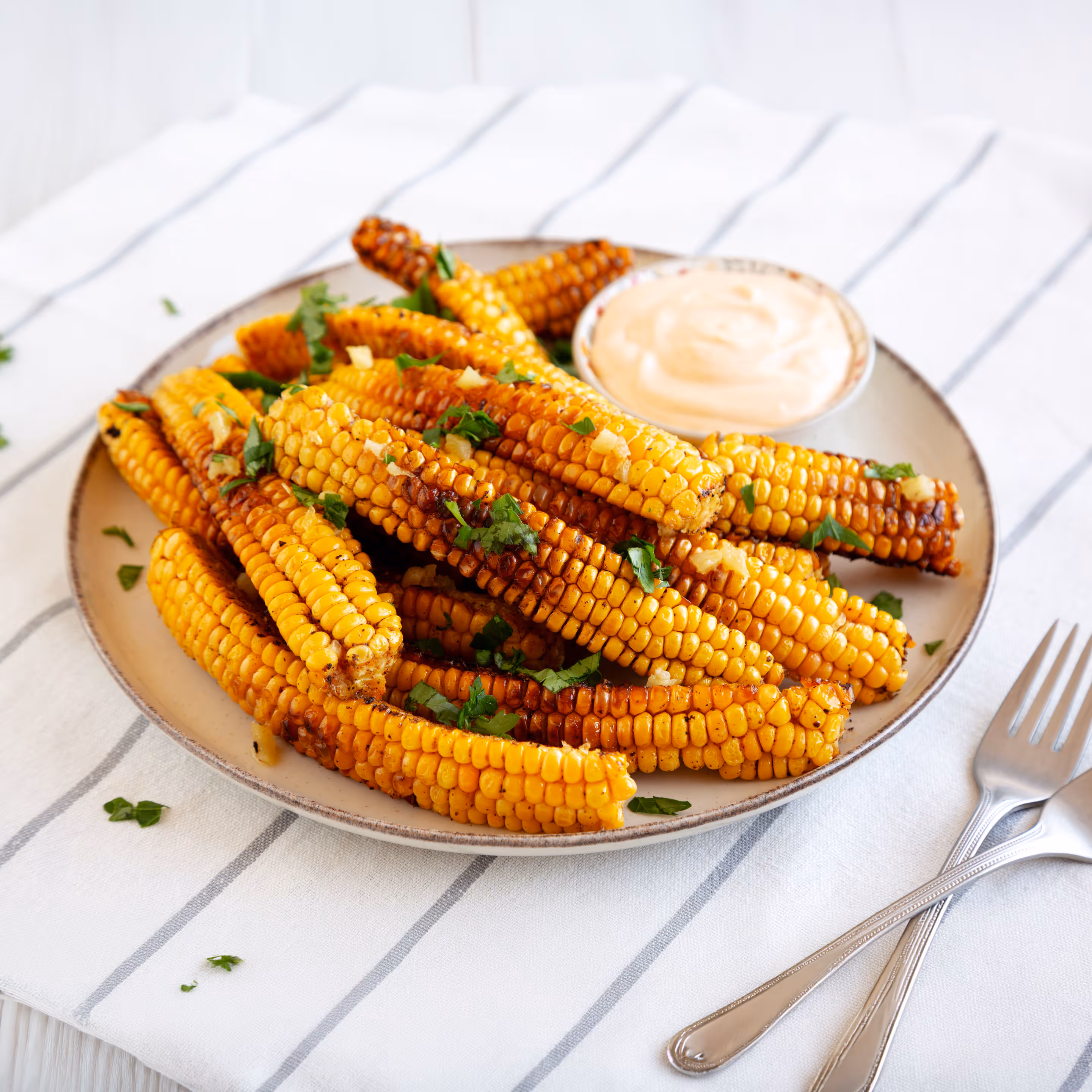 Grilled corn ribs sprinkled with chopped herbs on a plate with a creamy dipping sauce, set on a striped cloth with two forks nearby.