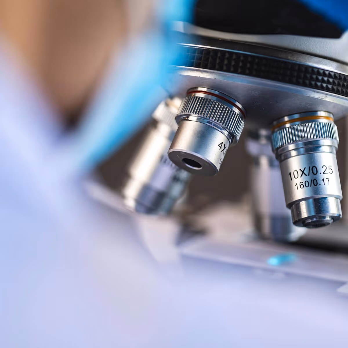 Close-up of microscope lenses labeled 4X and 10X with a blurred person wearing a blue mask in the background.