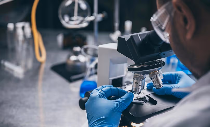 Scientist wearing blue gloves and protective eyewear adjusting a microscope slide in a laboratory.