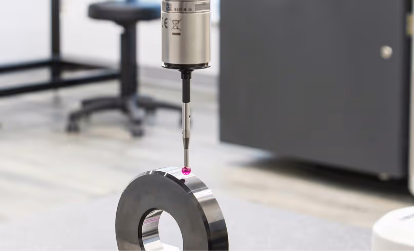 CMM probe with a red tip measuring a metallic circular object with a hole in the center on a workshop table.