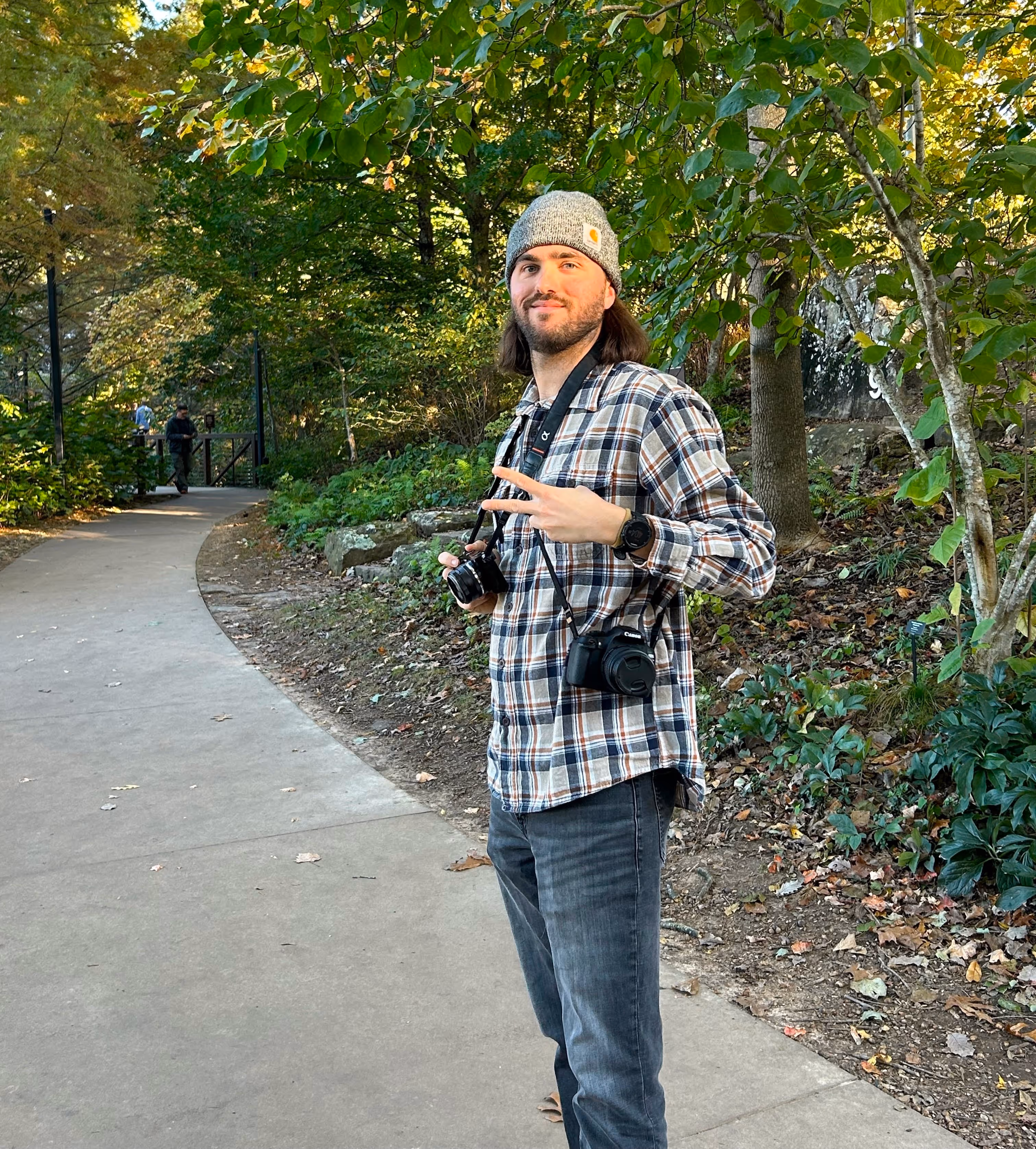 Man in a gray beanie and plaid shirt standing on a park pathway holding two cameras and making a peace sign.
