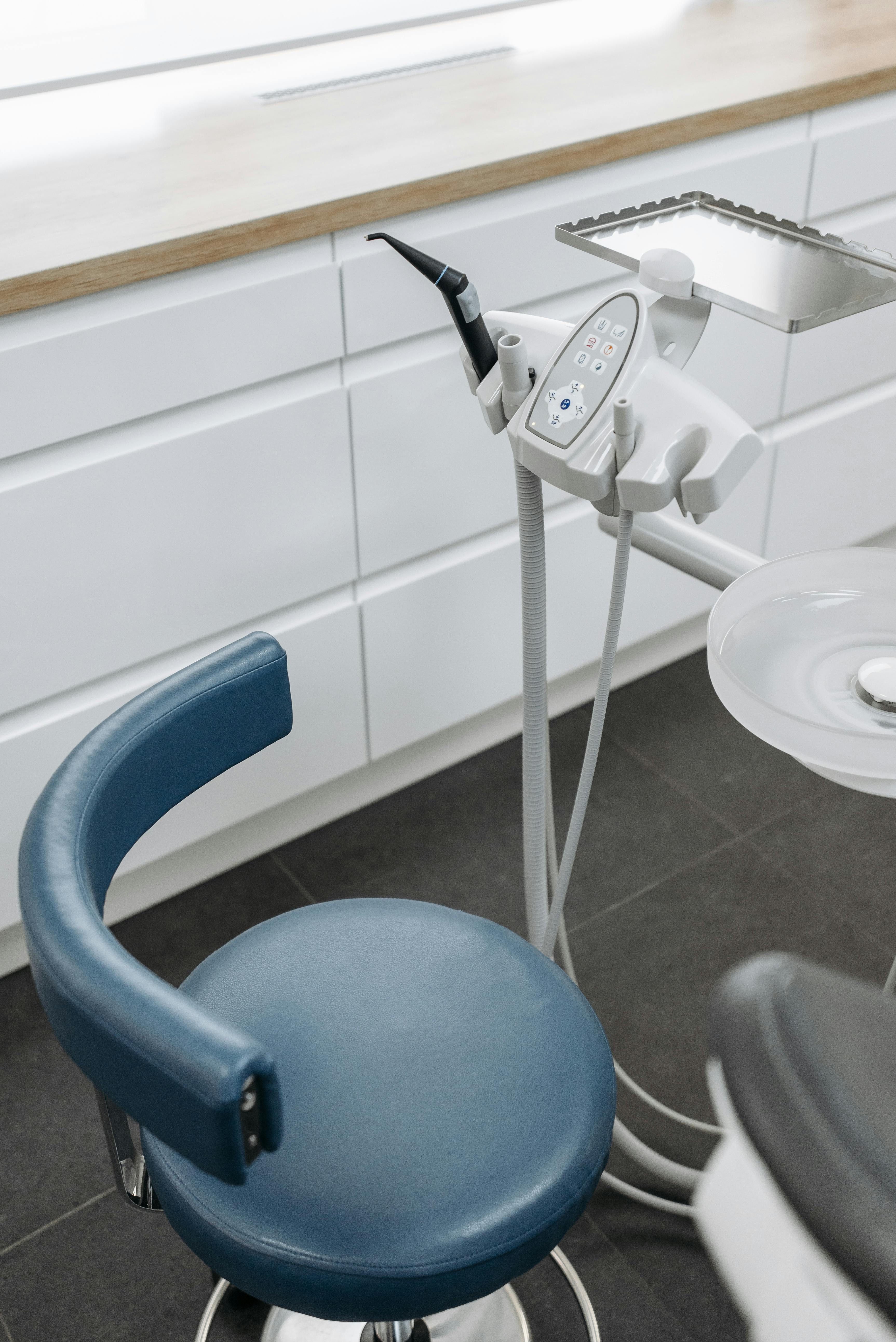 Blue cushioned dental stool next to a dental tool tray and spittoon in a dental clinic.