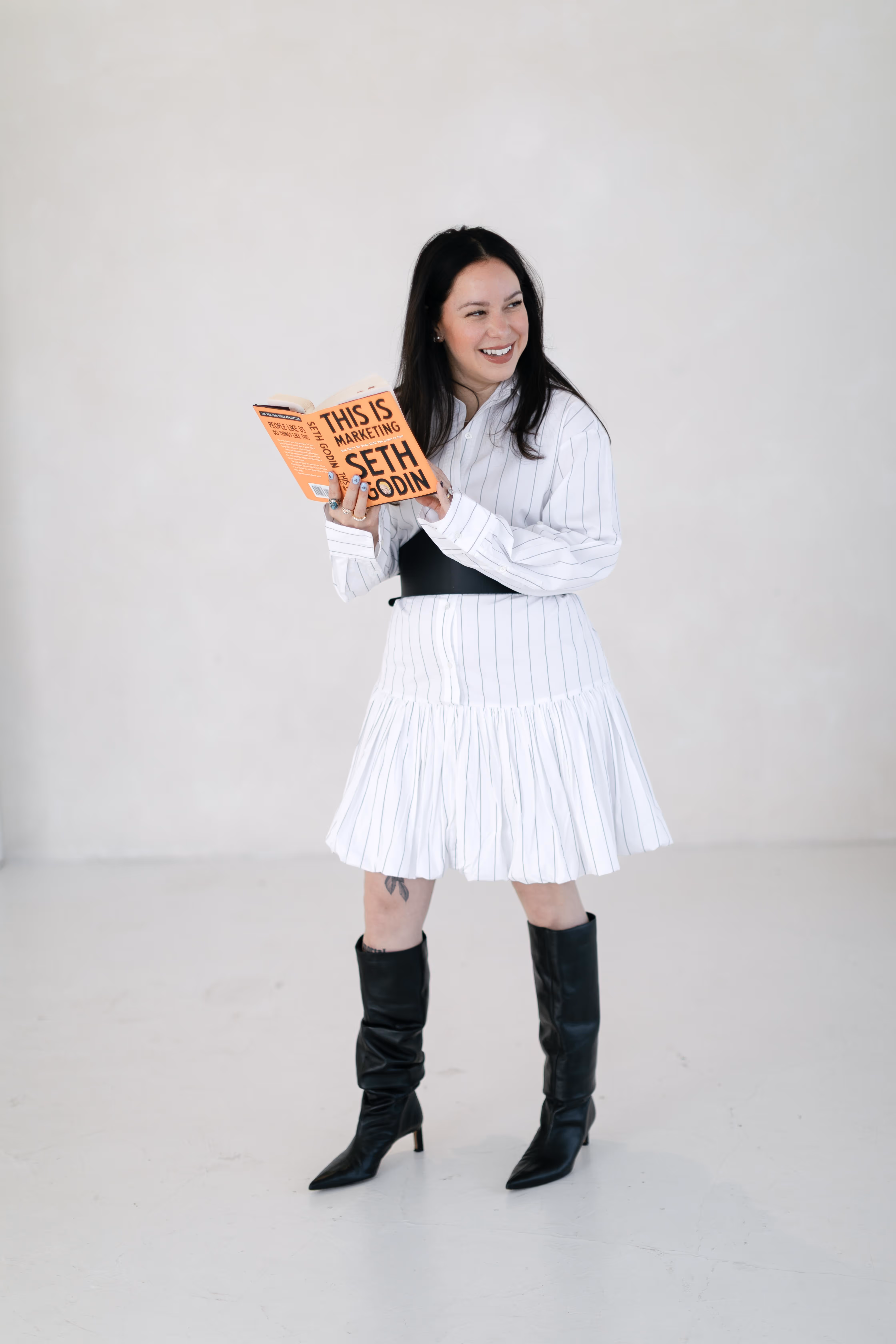 Woman in white dress and black boots smiling while holding an open book titled This Is Marketing.