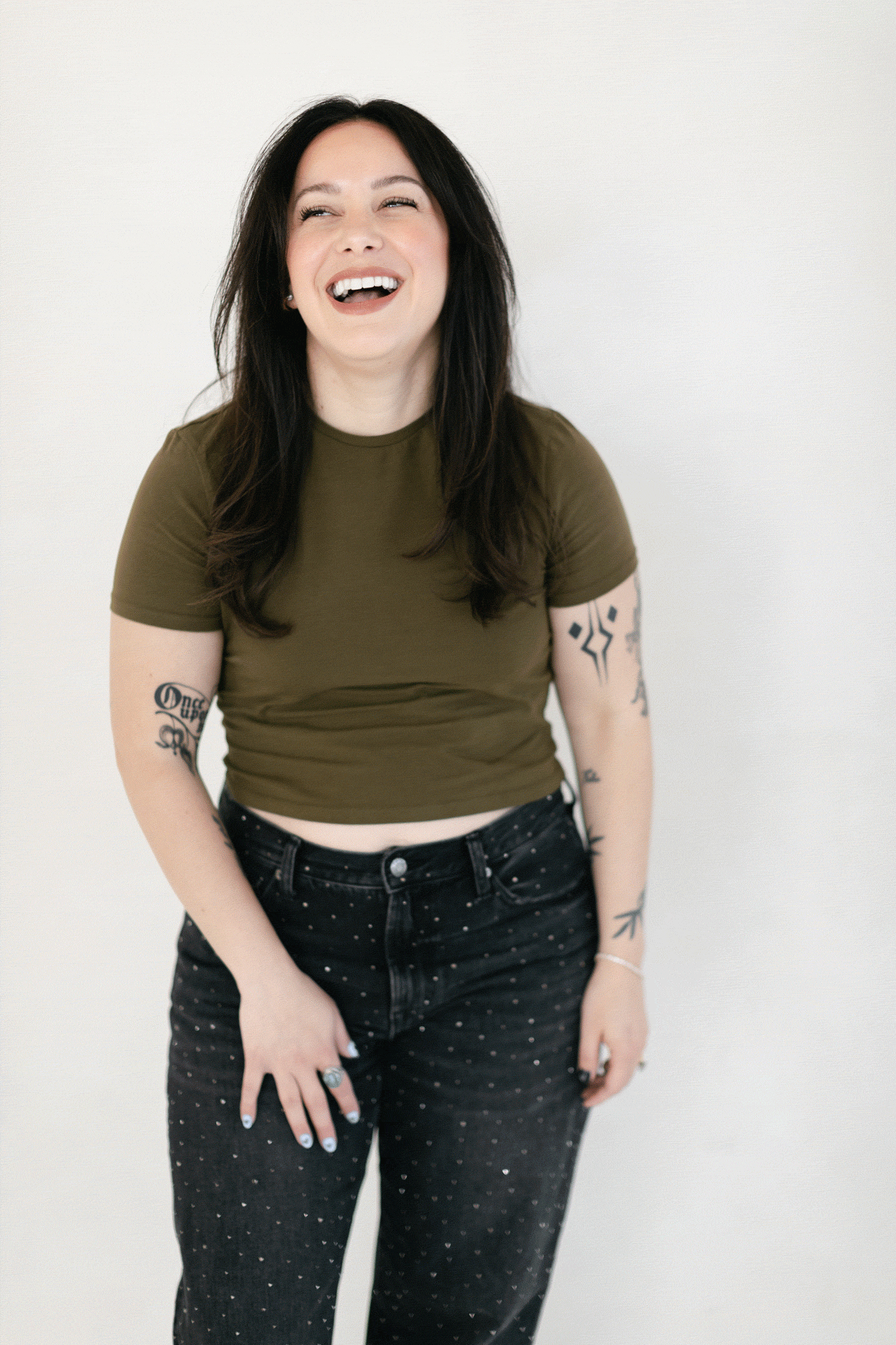 Young woman with tattoos laughing, wearing an olive green crop top and black polka dot jeans.